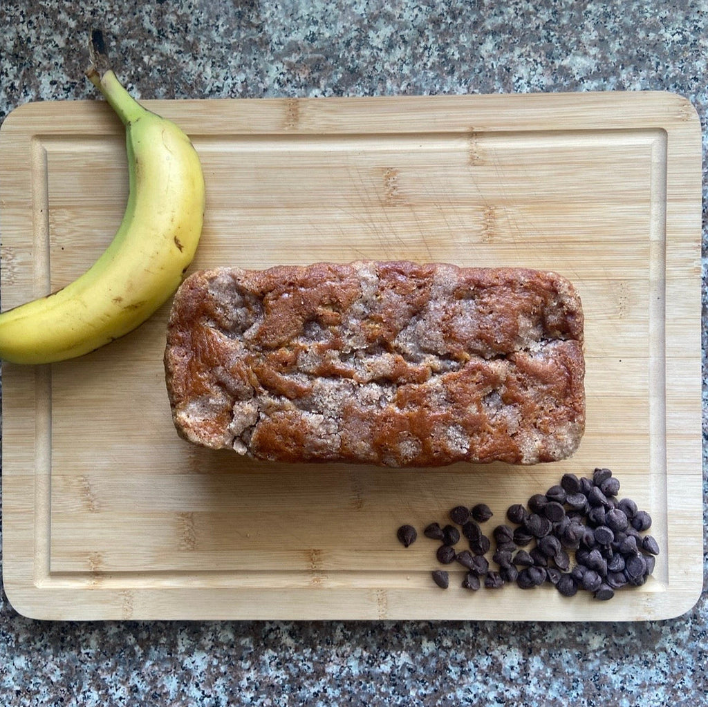 Loaf of banana bread with a banana and chocolate chips on a wooden cutting board.