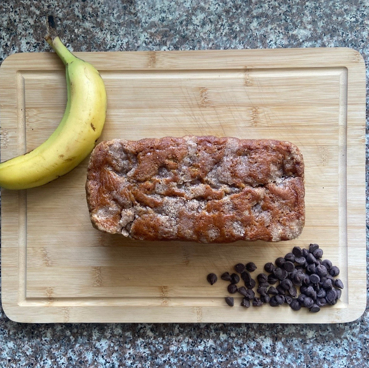 Loaf of banana bread with a banana and chocolate chips on a wooden cutting board.